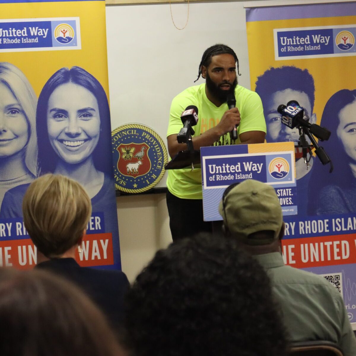 A Black man wearing a neon yellow T-shirt and speaking from a podium.